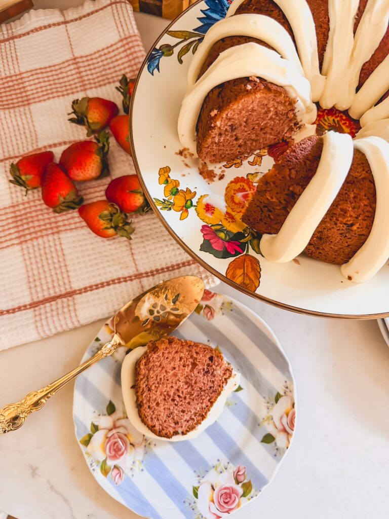 slice of strawberry bundt cake on flower plate with gold cake server. whole bundt cake on serving platter next to it with strawberries on dish towel.