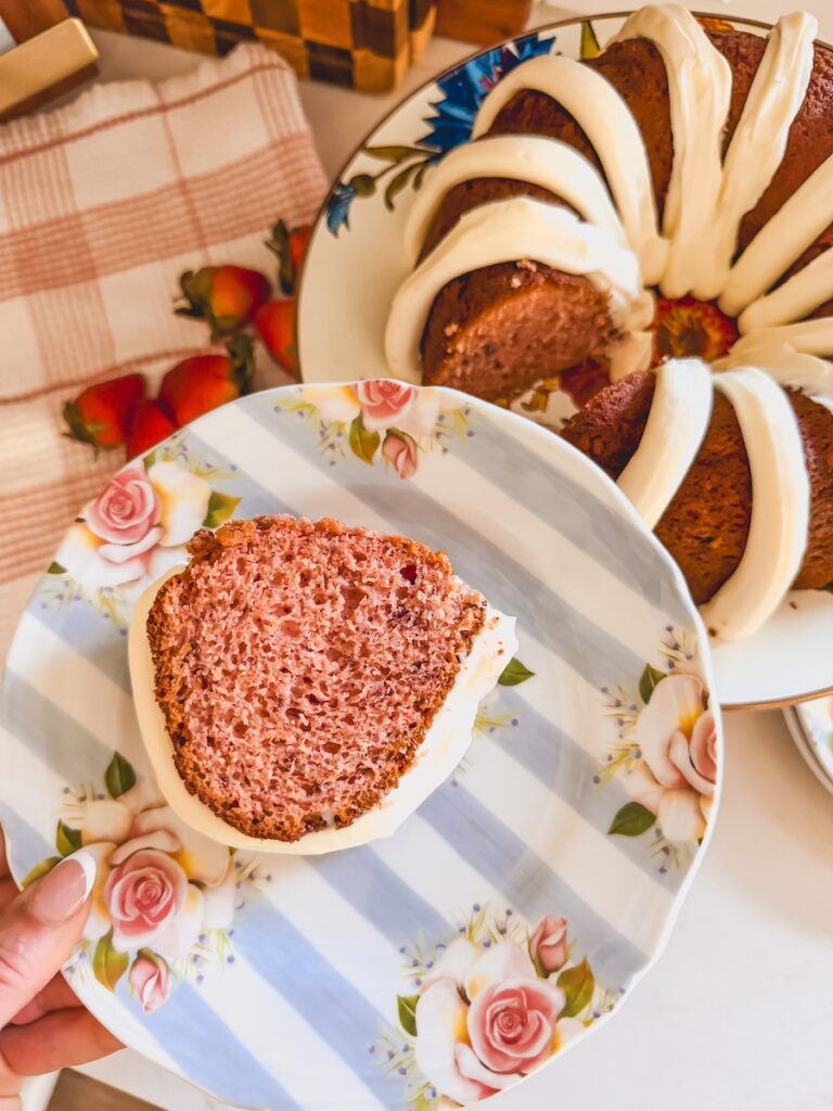 hand holding flower plate with slice of strawberry bundt cake on it. whole bundt cake in background.