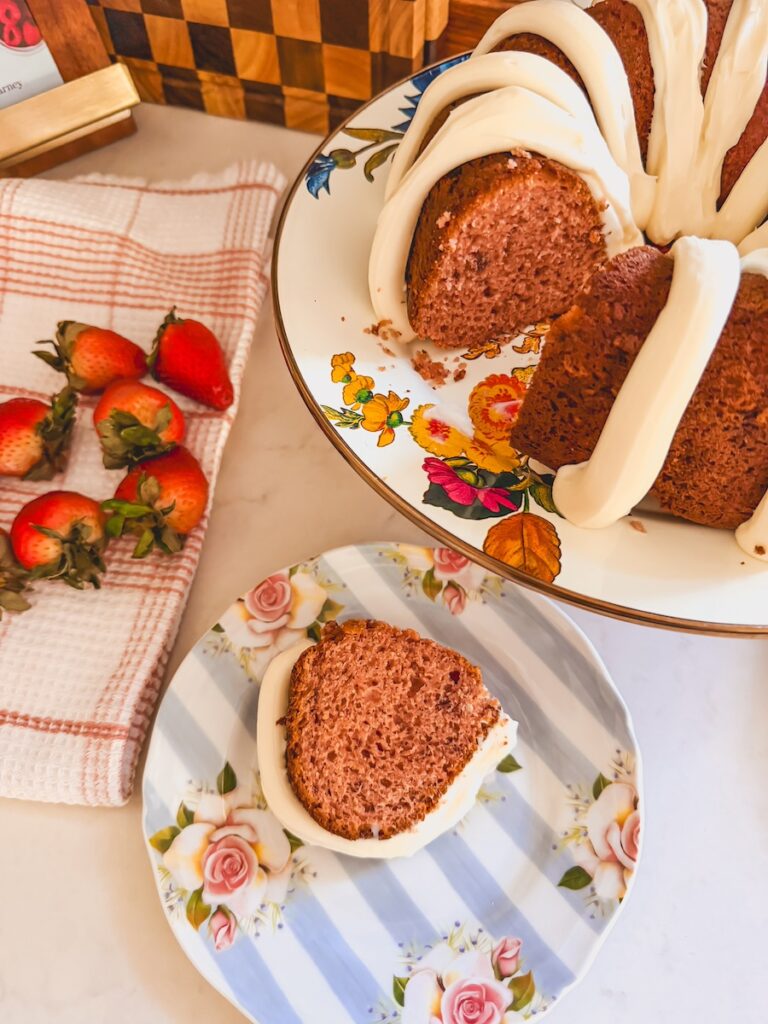 slice of strawberry bundt cake on flower plate next to whole strawberry bundt cake and fresh strawberries