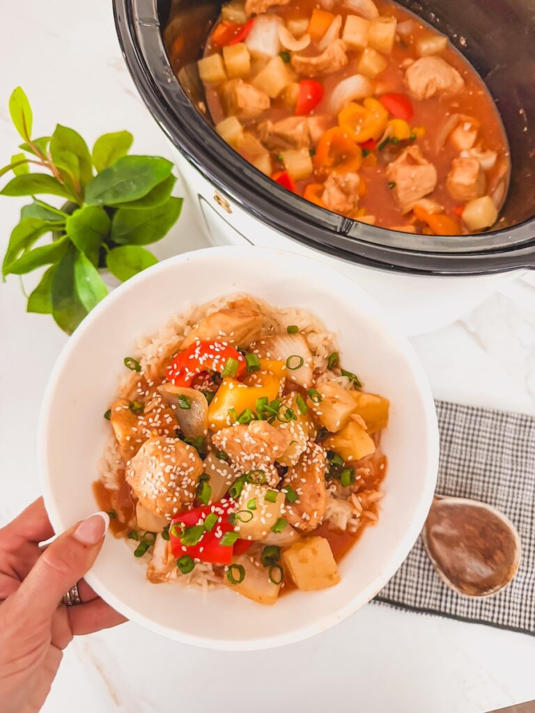 hand holding bowl of sweet and sour chicken with crockpot of sweet and sour chicken in background