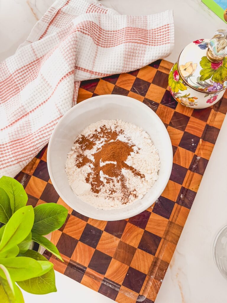 A white bowl with dry ingredients for carrot banana muffins on a checkered cutting board.