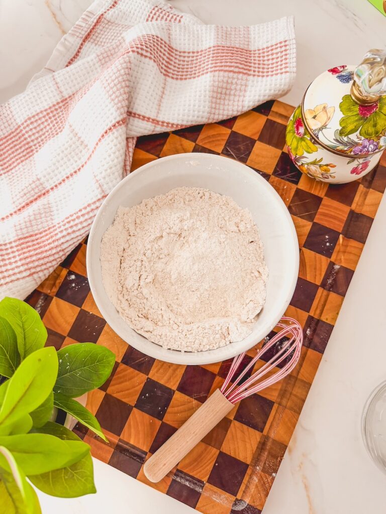 A white bowl with dry ingredients and a whisk to the right side on a checkered cutting board. A linen is on the left side.