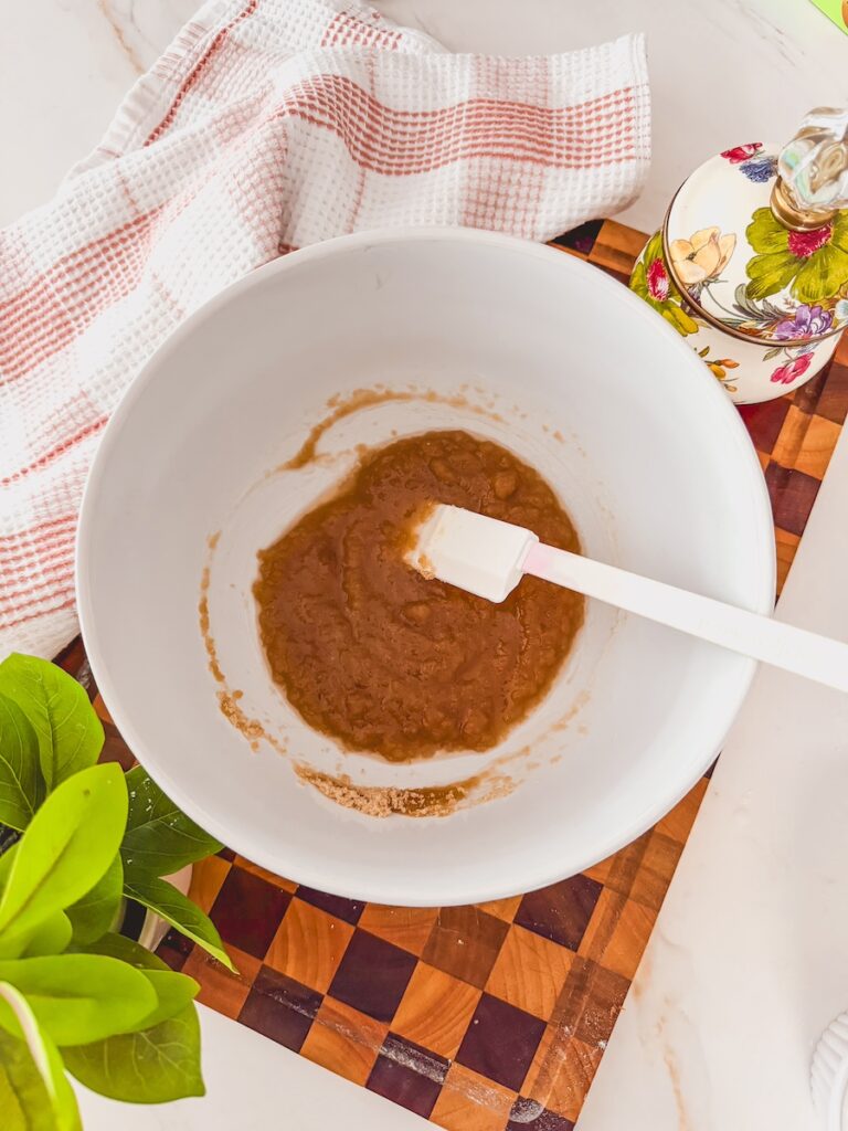 A white bowl with wet ingredients and a white spatula on a wooden cutting board. A linen is to the side.