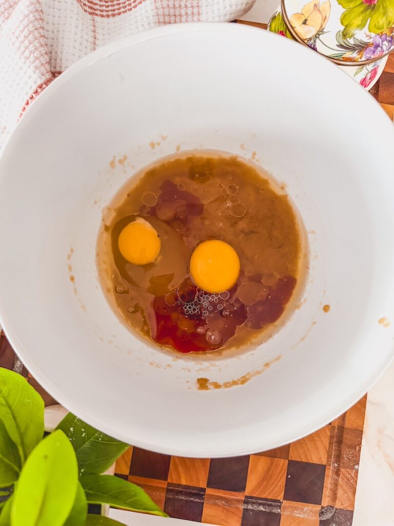 A white bowl with wet ingredients and two eggs cracked on top. The bowl is on a checkered cutting board.