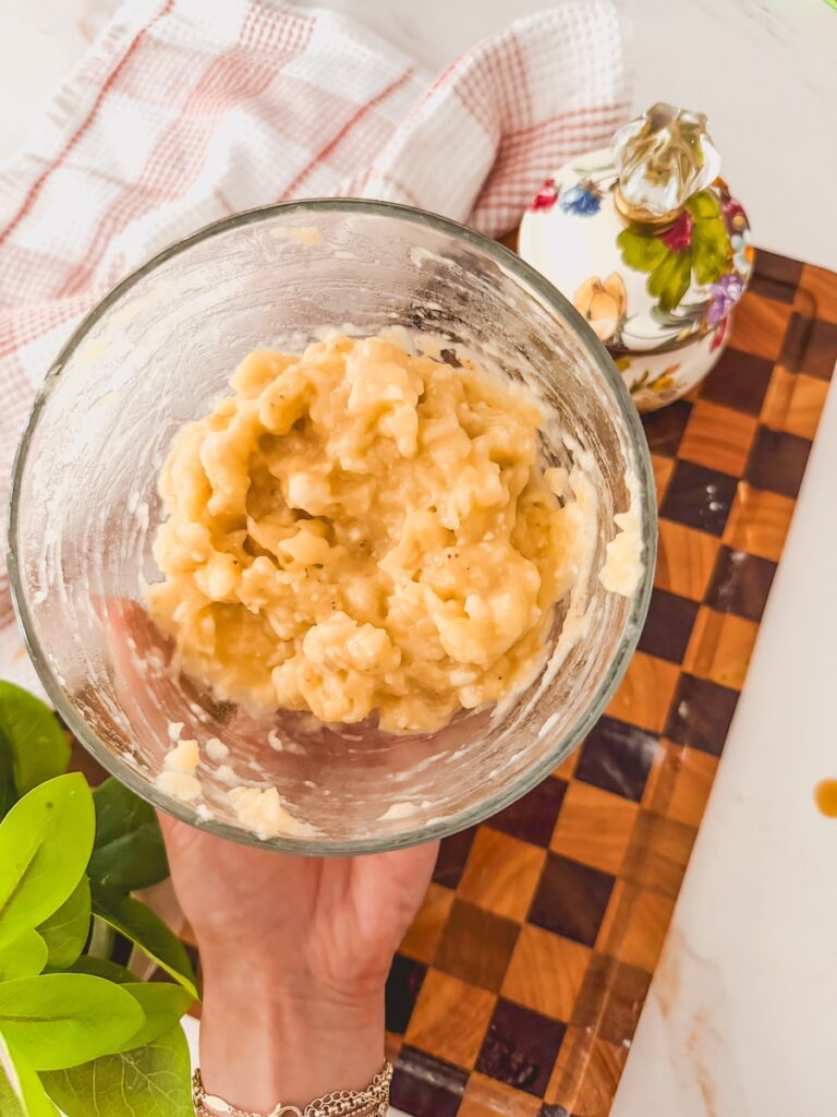 A glass bowl being held with a hand with mashed bananas. A checkered cutting board is in the background.
