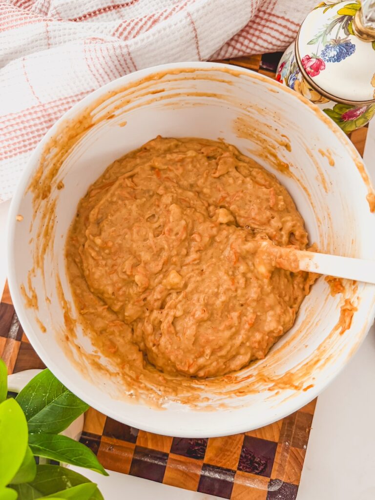 A white bowl with carrot banana muffin batter with a white spatula resting on the side.