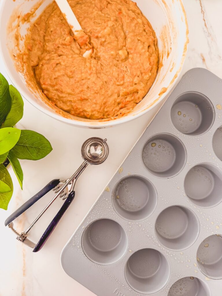 A white bowl with carrot banana muffin batter with a white spatula resting in the bowl. A muffin tin is to the left of the bowl with a cookie scoop in the center.