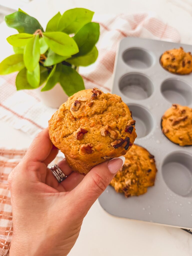 A hand holding carrot banana muffins with more in a tin in the background.