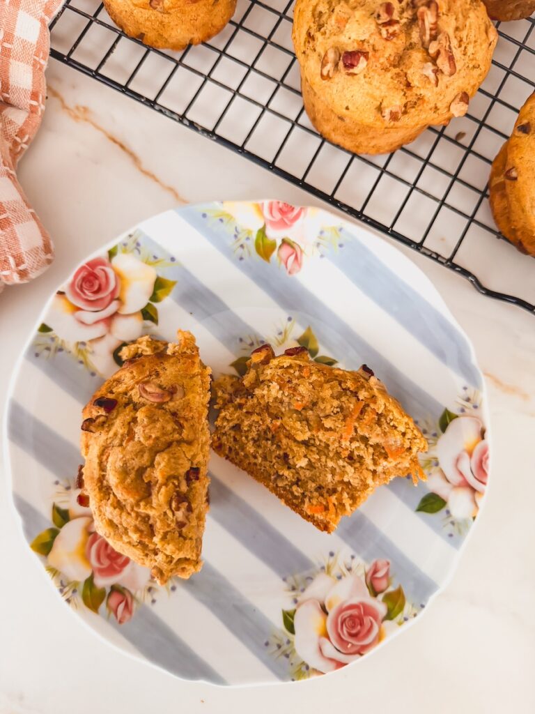A carrot banana muffin is cut in half on a flower plate, others are cooling on a wire rack.