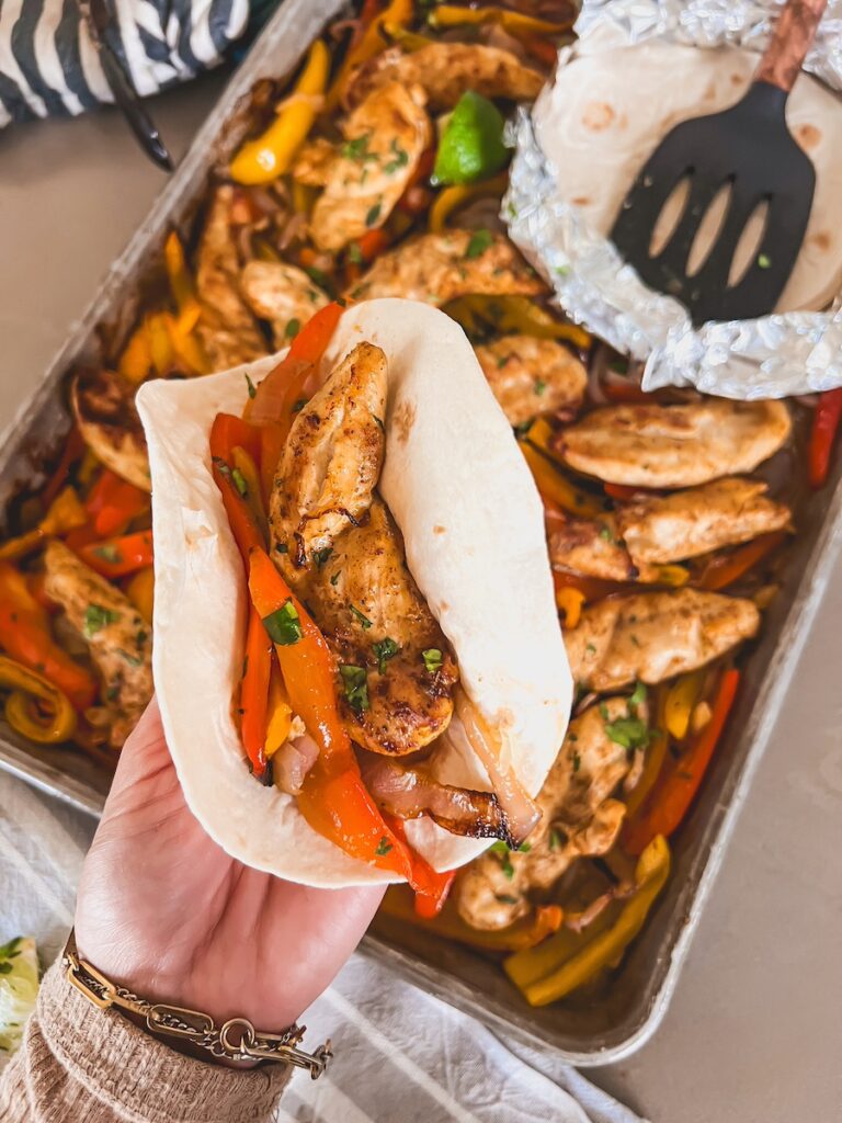 Hand holding a tortilla with chicken fajitas and sheet pan with fajitas on it in the background