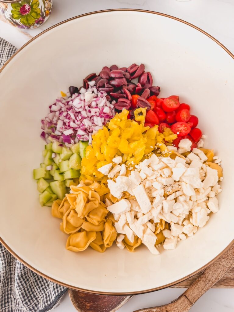 greek tortellini salad components in white bowl ready to be combined