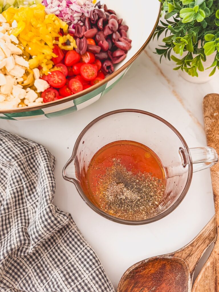 greek tortellini salad dressing in glass measuring cup ready to be mixed with salad ingredients in bowl next to it, also ready to be mixed