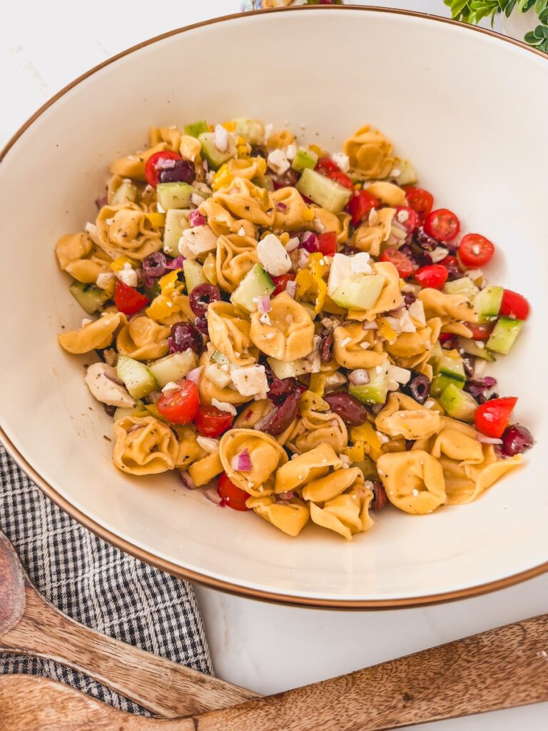 another overhead-side view of Greek Tortellini Salad in white bowl. dish towel and serving spoons next to bowl.