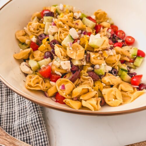 another side view of Greek Tortellini Salad in white bowl. dish towel and serving spoons next to bowl.