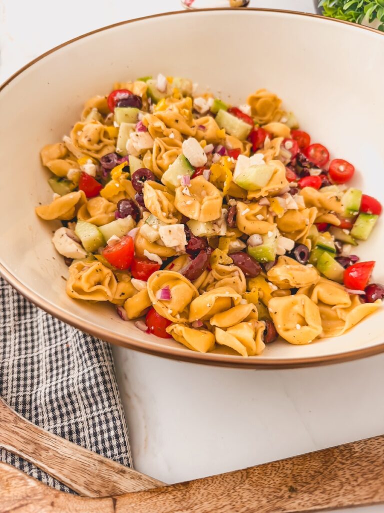 another side view of Greek Tortellini Salad in white bowl. dish towel and serving spoons next to bowl.