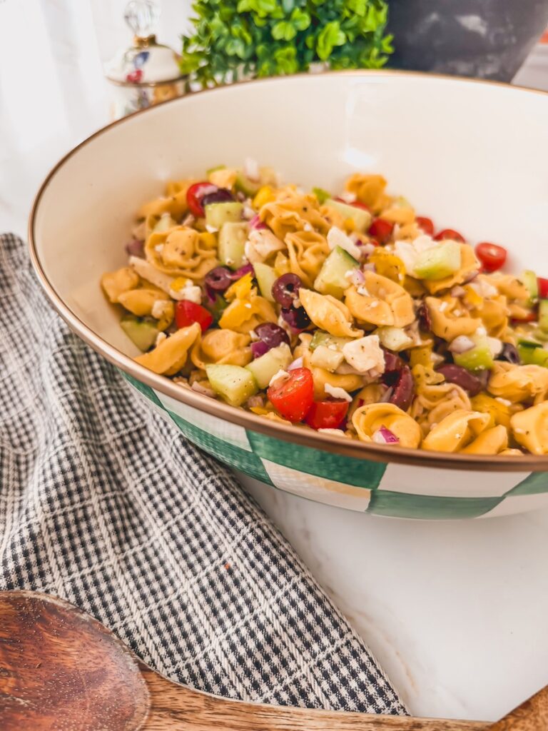 side view of Greek Tortellini Salad in white bowl with dish towel and serving spoons next to bowl.