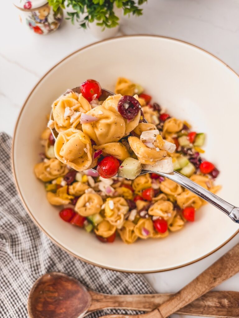 Greek Tortellini Salad in white bowl with spoonful of salad. dish towel and serving spoons in background.