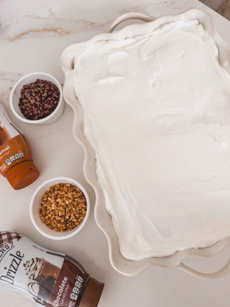 another layer of whipped topping added to the ice cream cake. Chopped nuts, chocolate chips, fudge and peanut butter sauce bottles next to ice cream cake on counter.