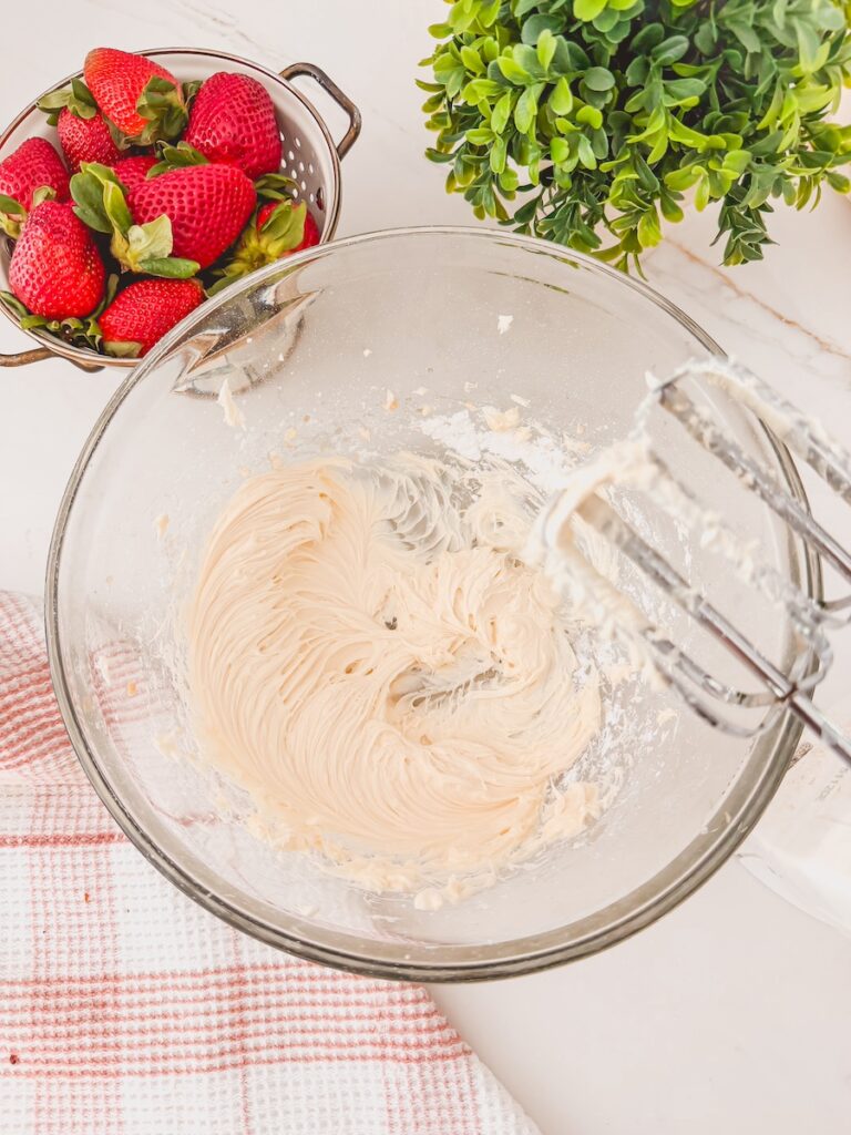 handmixer whipping pudding filling in glass bowl. whole strawberries in colander and greens in background.