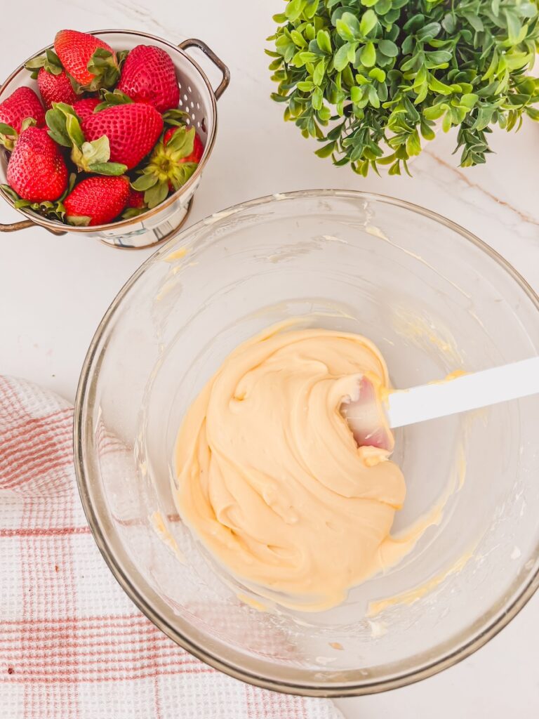 pudding mixture in glass bowl with spatula. whole strawberries in colander and greens in the background.