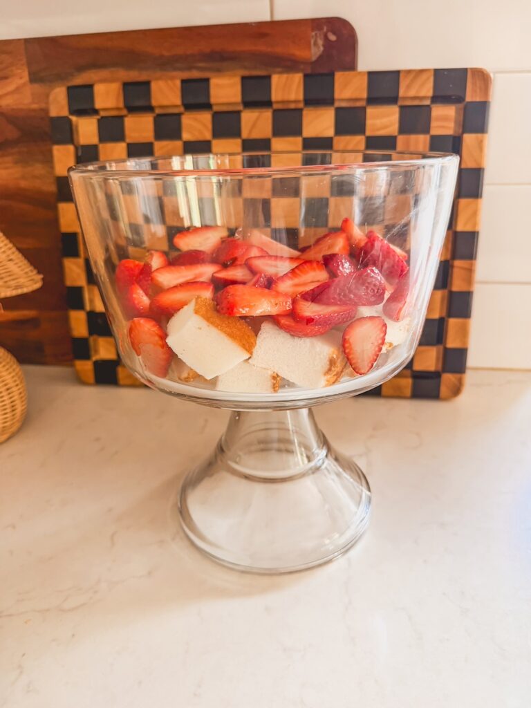 strawberry shortcake trifle being built in glass trifle dish on counter with cubed cake and sliced strawberries.