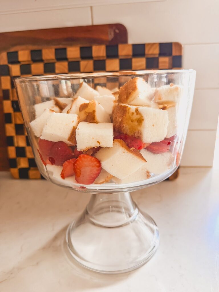 strawberry shortcake trifle being built in glass trifle dish on counter with cubed cake and sliced strawberries, plus another layer of cubed cake.