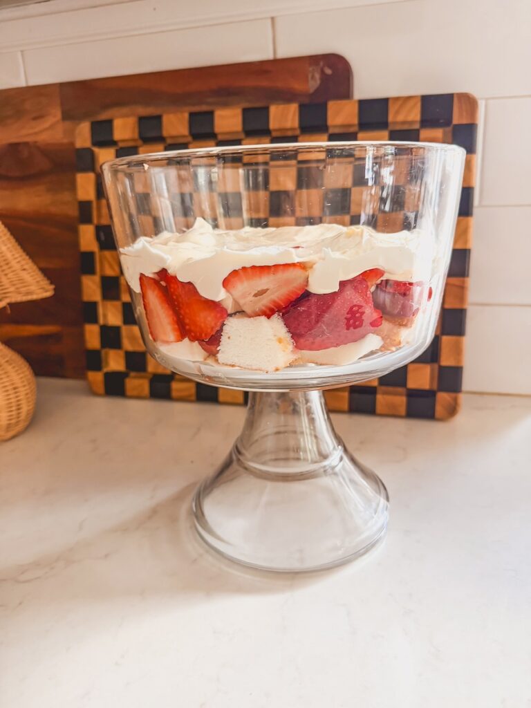 strawberry shortcake trifle being built in glass trifle on counter with cubed cake, sliced strawberries and pudding filling. checked cutting board in background.