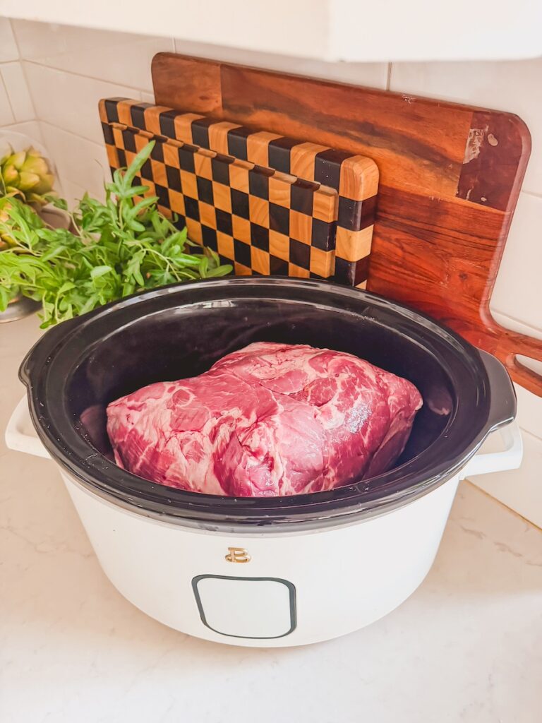 Raw pork in slow cooker on counter. cutting boards in background.
