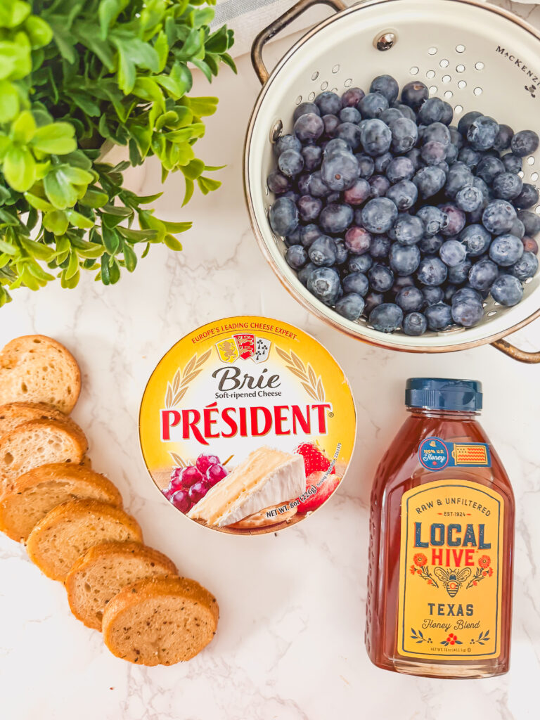 Ingredients for baked blueberry brie with honey including brie cheese, fresh blueberries, and honey on white background