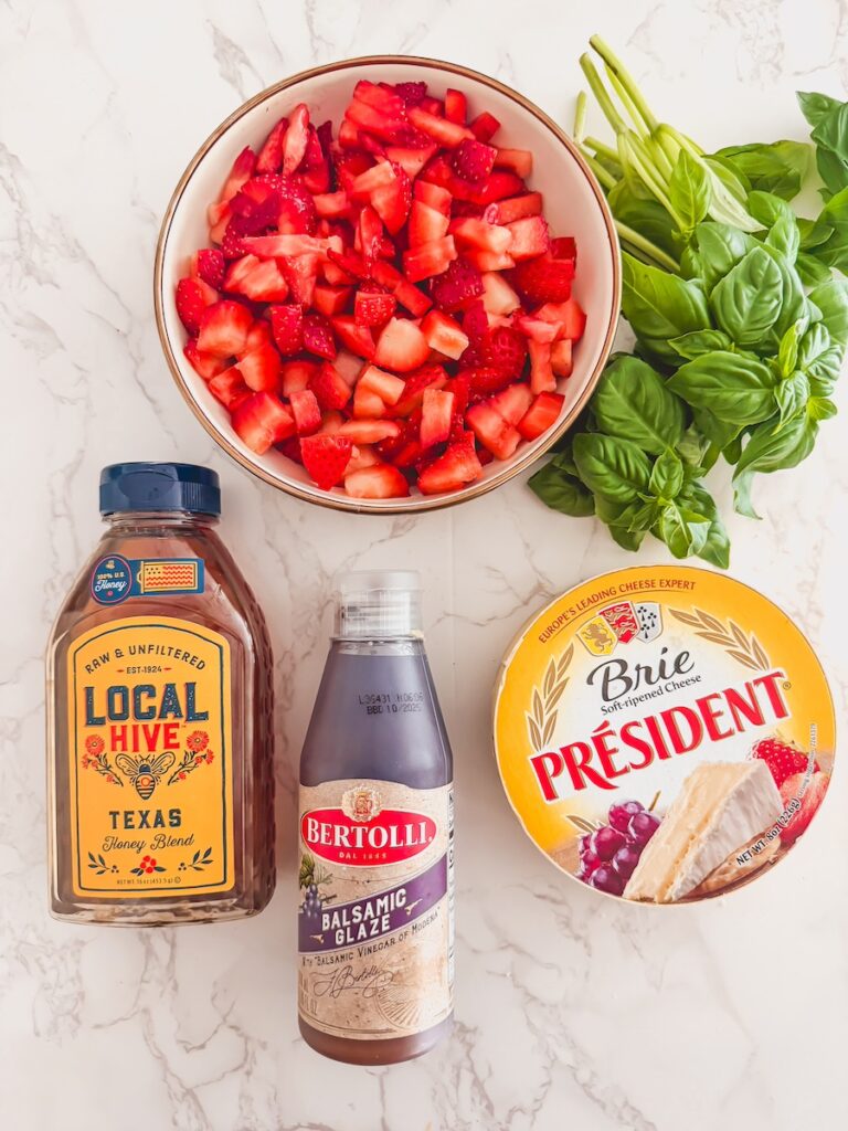 flatlay of ingredients for strawberry baked brie with basil, strawberries, brie, honey and balsamic glaze