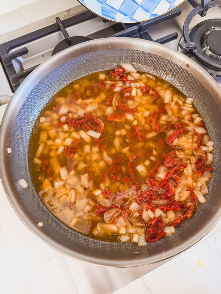 A skillet with broth, onion, and sundried tomatoes.