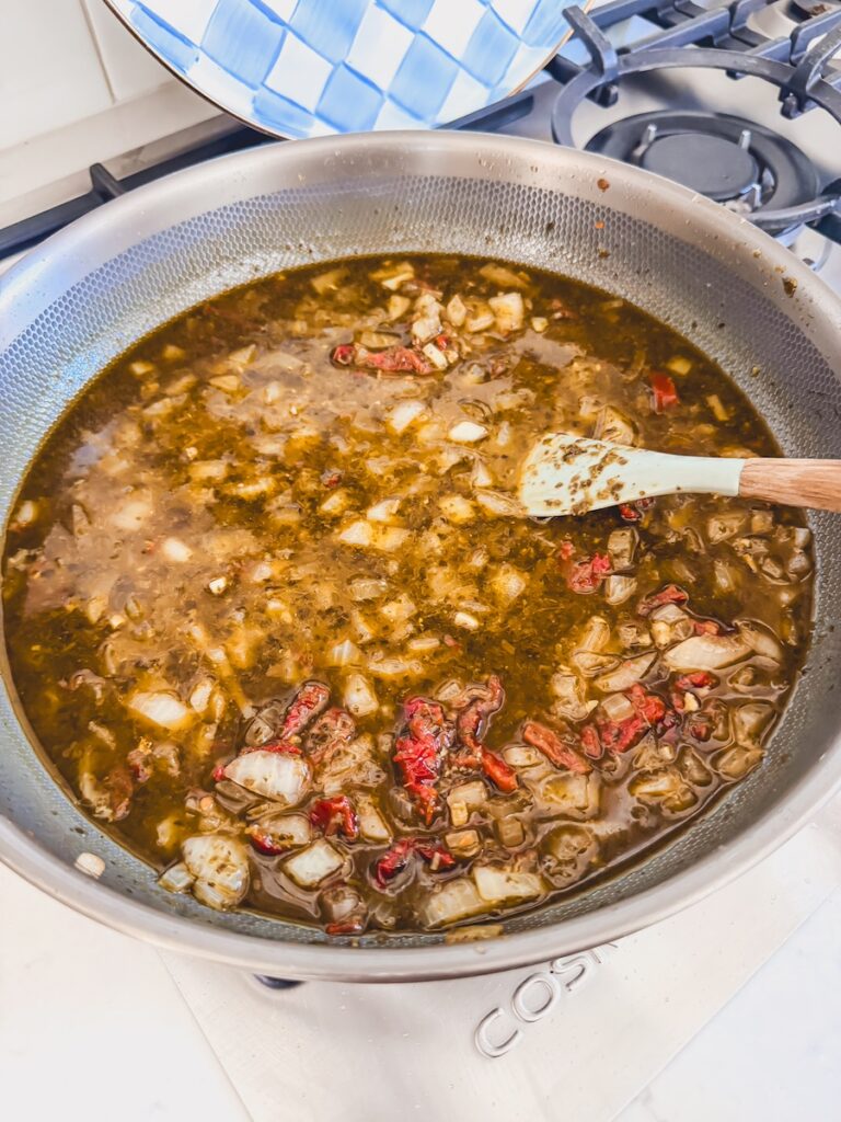 A skillet with broth, spices, onions, and sundried tomatoes.