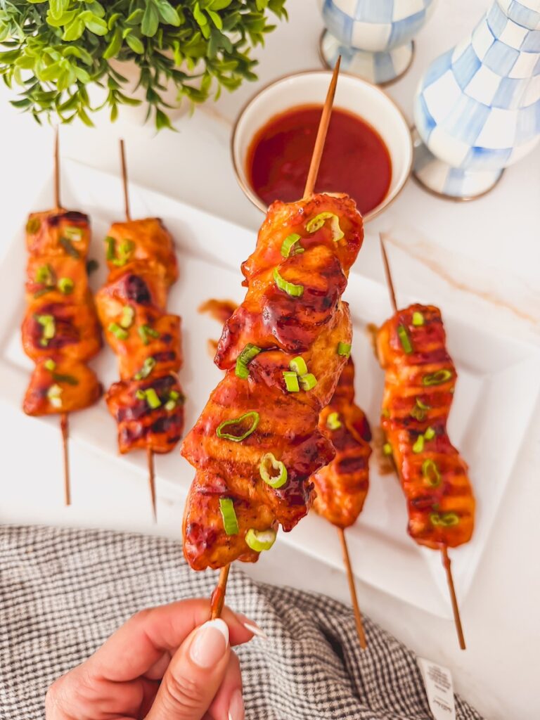 A hand holding a grilled bbq chicken skewer over a white platter of additional skewers.