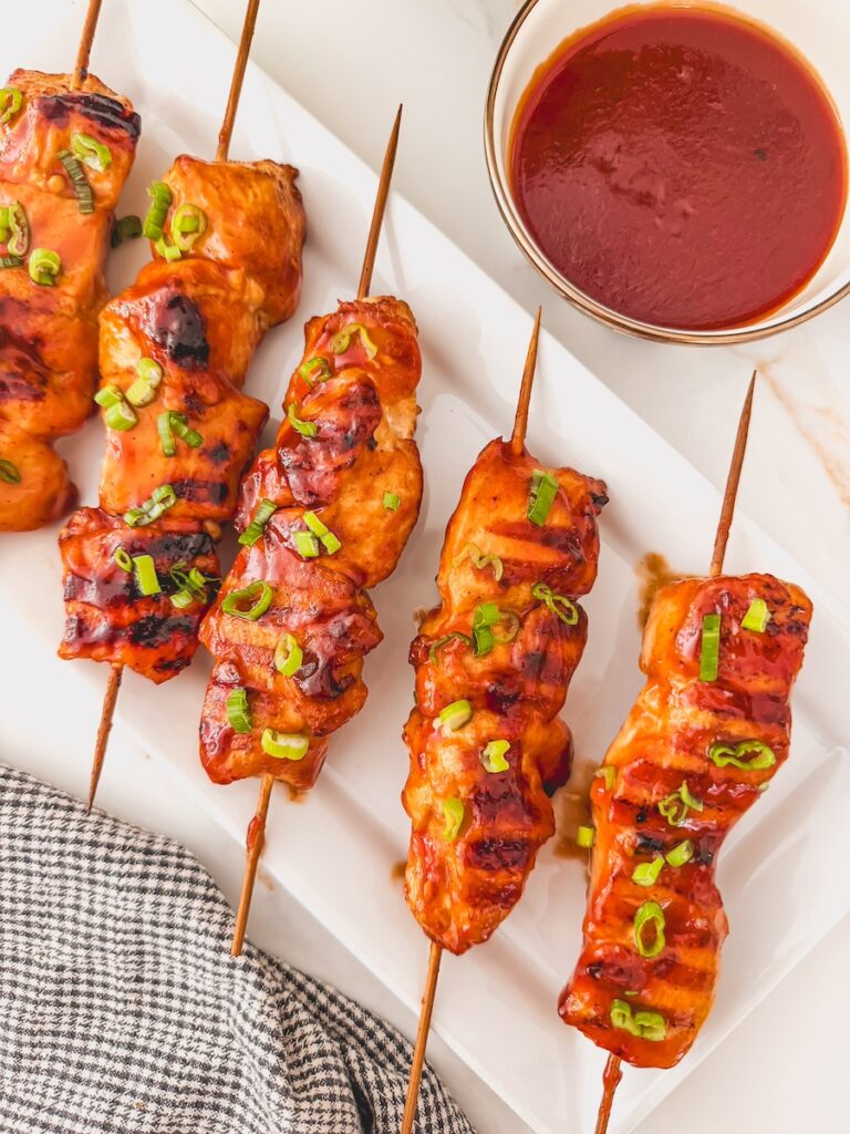 Overhead image of bbq chicken skewers on a white plate with a glass bowl of homemade bbq sauce on the side.