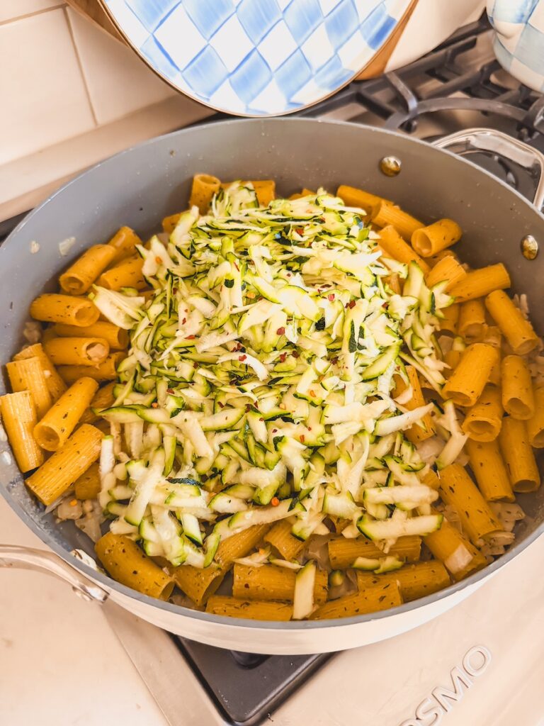 A pan with noodles, onion, garlic, grated zucchini and chili flakes.