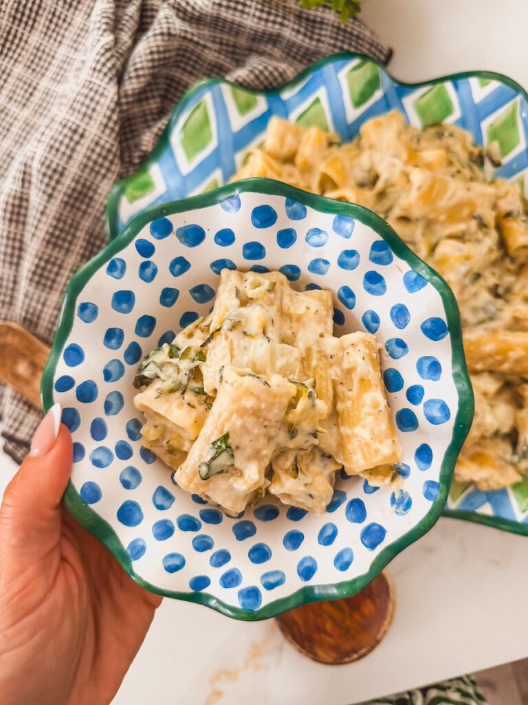 A blue polka dot bowl with a green rim of lemon zucchini pasta and a hand holding it.