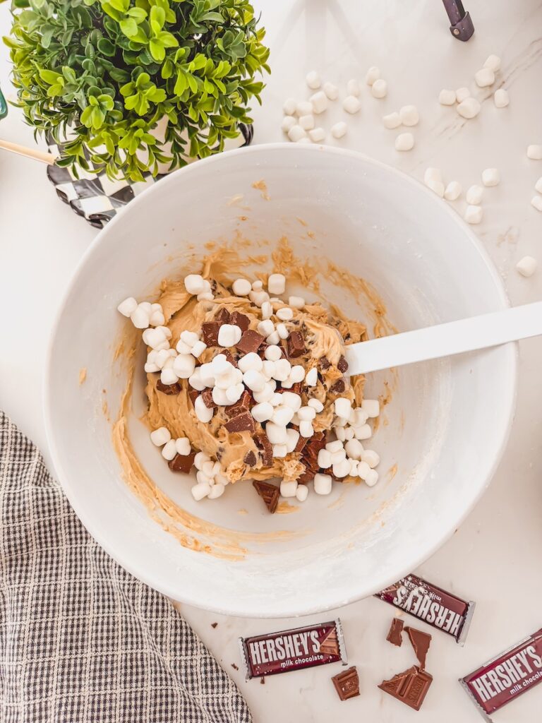 A large white bowl with cookie dough and a white spatula stirring in marshmallows and chocolate chunks.
