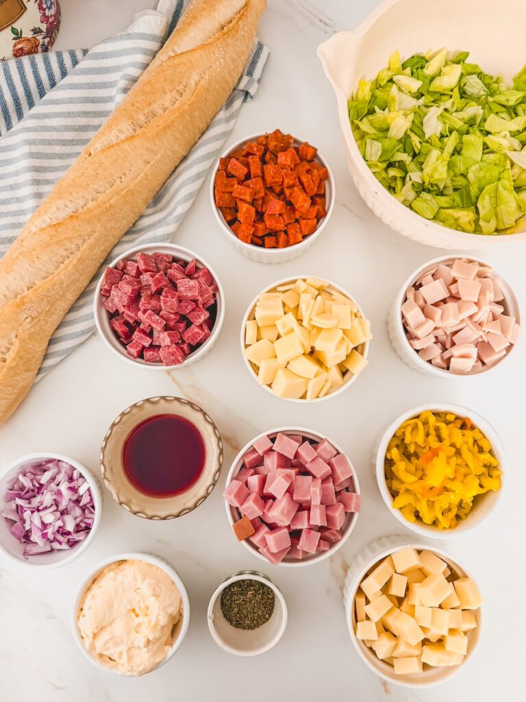 flatlay of ingredients for italian hoagie dip - lettuce, deli meats and cheeses, banana peppers, red onion, red wine vinegar, Italian seasoning and mayo