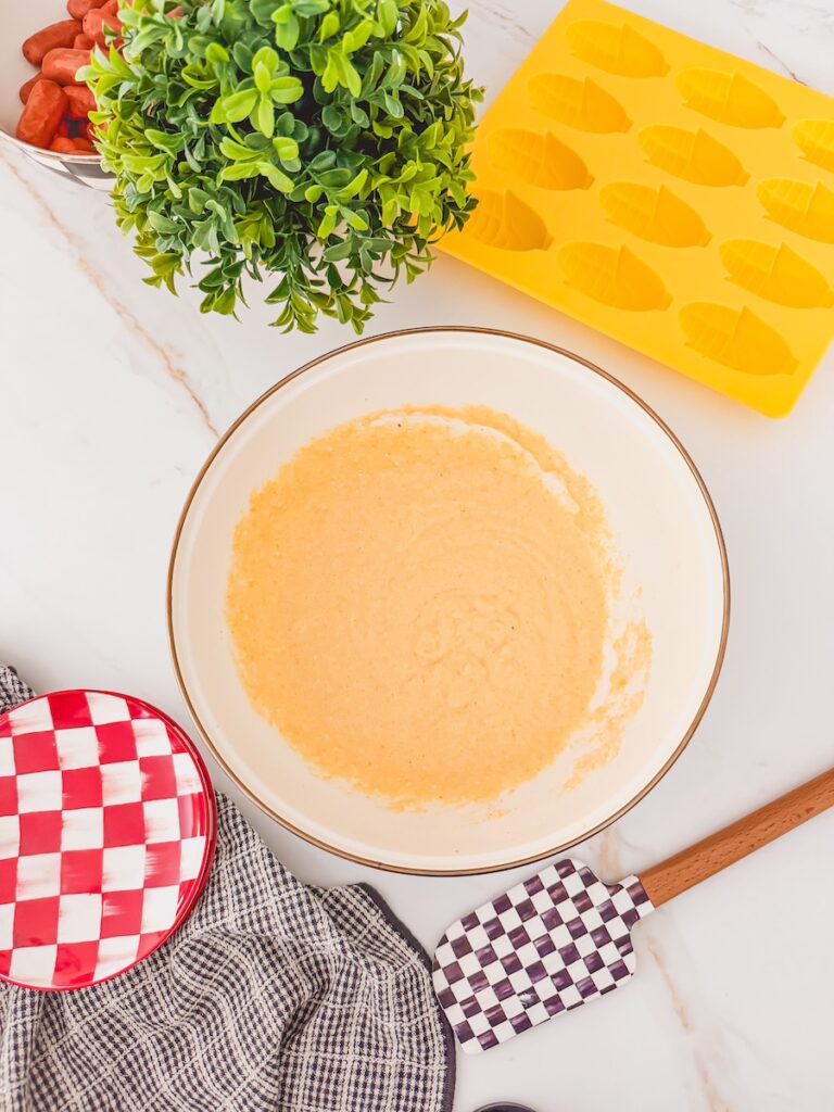 white bowl of corn dog batter with empty mold and side dishes on either side