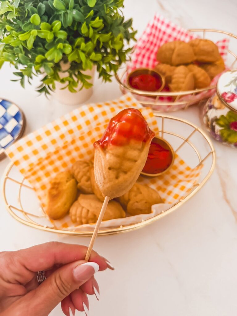hand holding finished homemade corn dog on skewer dipped in ketchup with baskets of corndogs in background