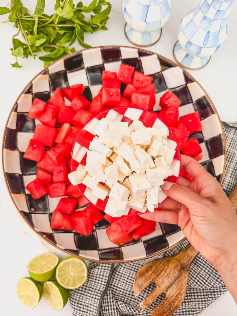 cubed watermelon in serving bowl with hand holding smaller bowl of feta cubes, ready to add in