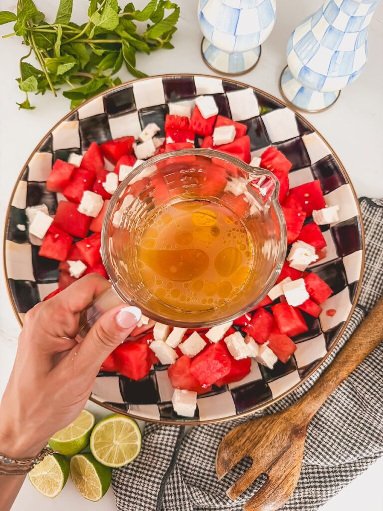 hand holding measuring cup of dressing with watermelon salad in serving bowl underneath it.