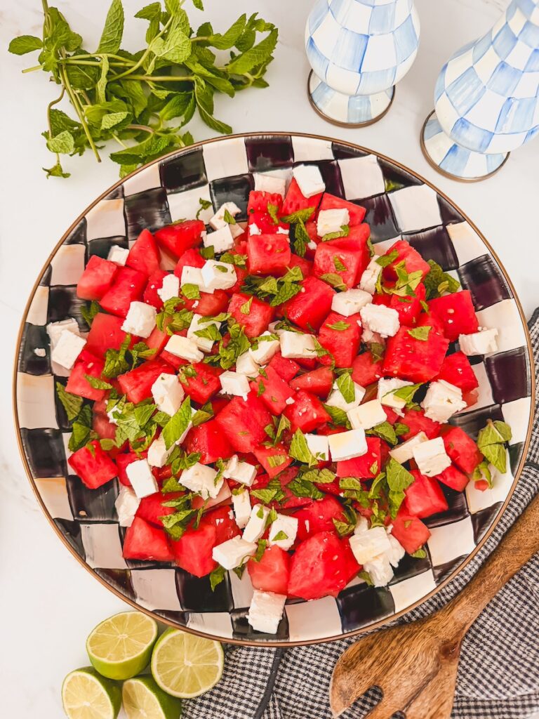 overhead image of finished and plated watermelon feta mint salad in black and white checked serving bowl with salt and pepper shakers, fresh mint, limes and wooden serving spoon next to it.