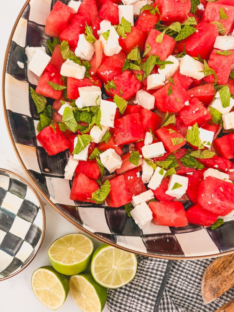 Finished and plated watermelon feta mint salad in black and white checked serving bowl with lime halves on side of bowl.