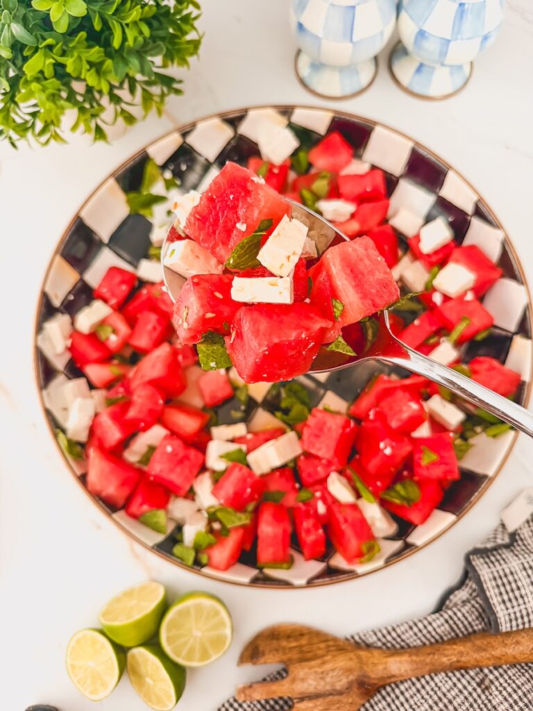 finished and plated watermelon feta mint salad in black and white checked serving bowl with spoonful of salad above. Limes and wooden serving spoon on counter.