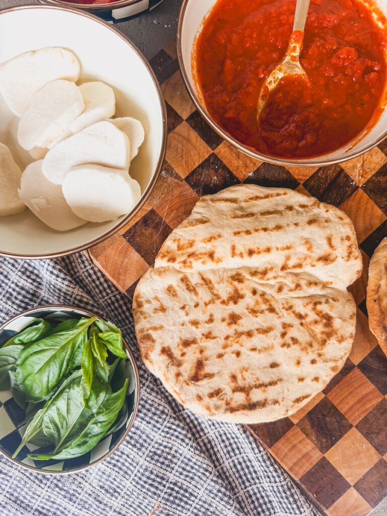 Assembly ingredients for grilled pizza which include grilled crust, marinara sauce, mozzarella slices, and fresh basil leaves. The ingredients are on a checkered cutting board with a checkered linen underneath.