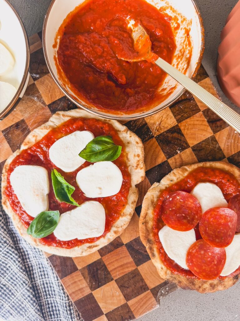The second pizza is being build with sauce, cheese, and basil. A bowl of sauce and second grilled pizza are next to it.