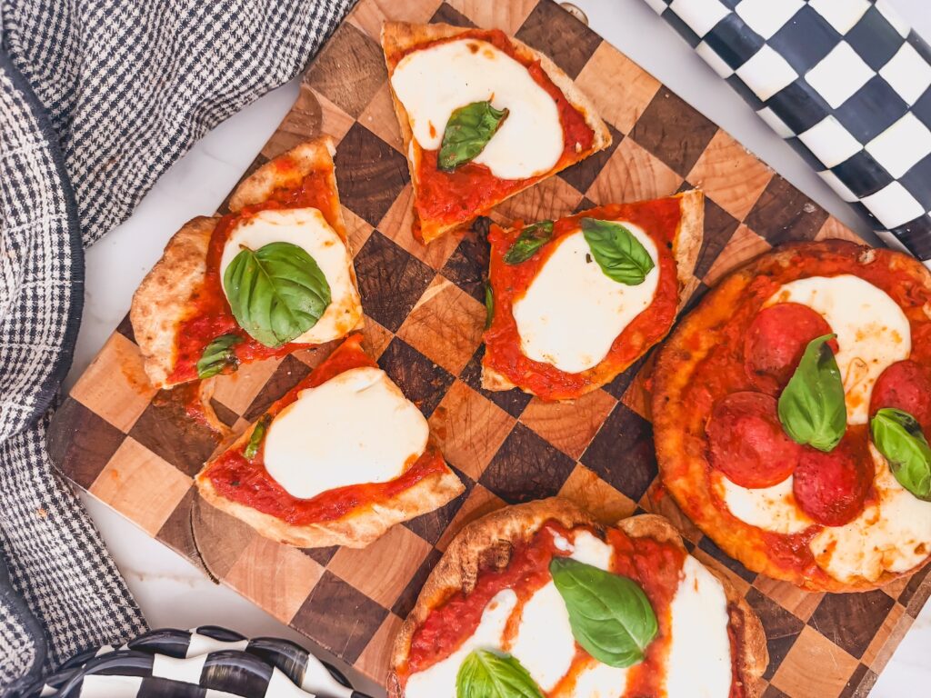 Grilled pizza is cut into four pieces with two whole grilled pizzas set to the side. The pizzas are on a checkered wooden cutting board.