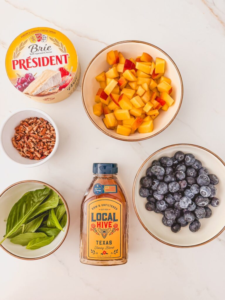 flatlay of ingredients; peaches, blueberries, brie, honey, basil and pecans