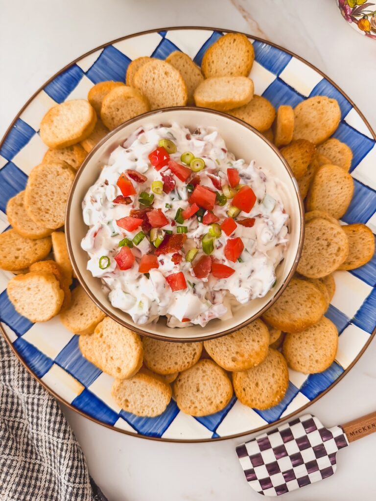 overhead view of bowl of blt dip in center of serving platter surrounded by crostini dippers. black and white checked spatula on the side.